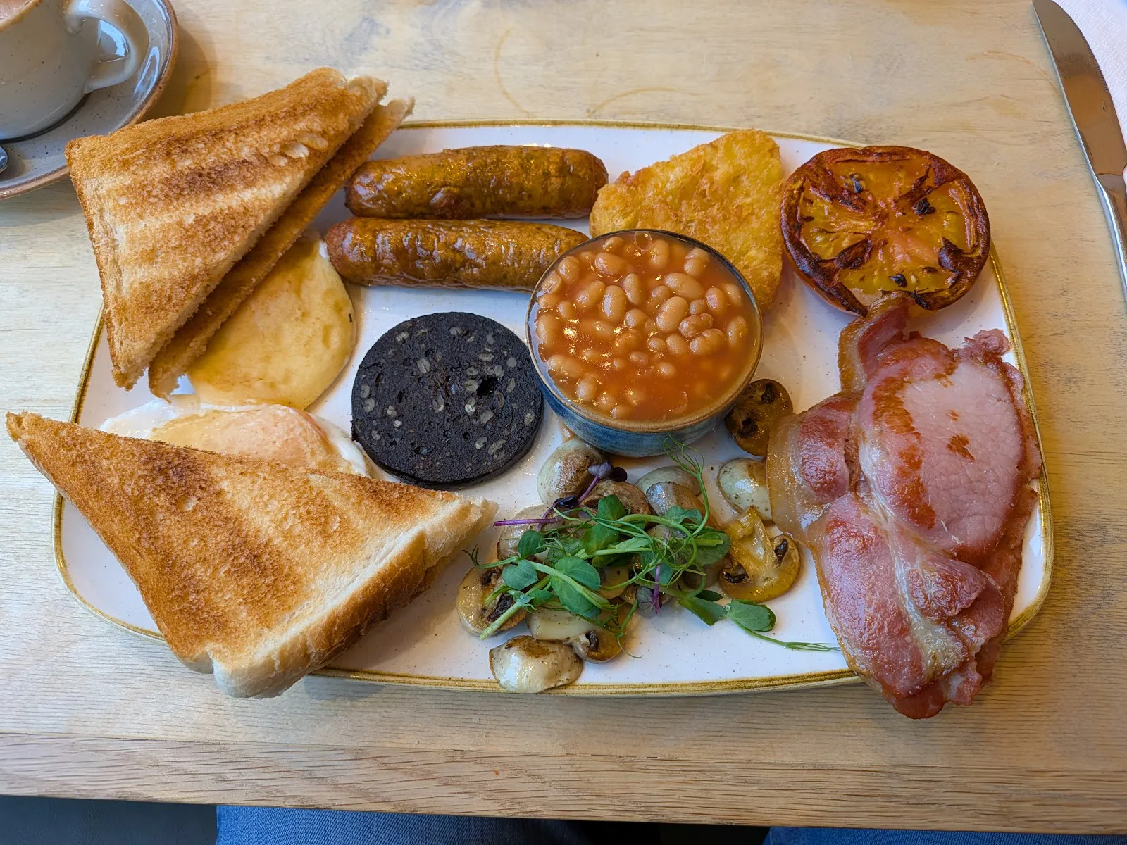 Full cooked breakfast on a gold-rimmed plate with toast, sausages, beans and bacon.
